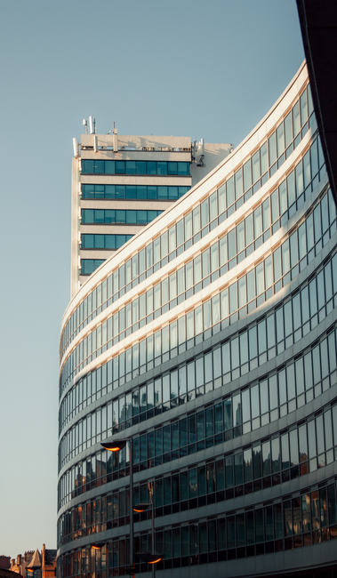 Photograph of a modern high-rise building with a curved glass facade reflecting the sky and surrounding structures. The building features multiple floors with large, tinted windows, framed by metallic trims, creating a sleek, contemporary appearance. The sunlight highlights the glossy surface, emphasizing cleanliness and shine. The sky is clear, providing a neutral backdrop, while streetlights at the base are visible. This image exemplifies urban architecture typical of commercial spaces, aligning with the theme of surface cleaning and maintenance. For professional deep cleaning and sanitisation services, iCarpet Cleaners ensures quality upkeep for such modern commercial properties.