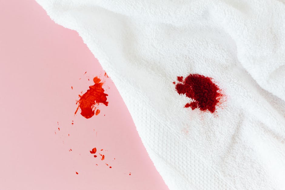 Close-up image showing a fresh red ink or blood stain on a white textured paper towel, contrasted with a light pink paper towel that has a similar but smaller stain. The paper towels are placed on a flat surface in a well-lit environment, emphasizing the contrast between the red stain and the white background. No cleaning tools or additional objects are visible in the image. The image is intended to depict the initial stage of a stain that may require emergency treatment, aligning with content about surface cleaning and stain removal procedures, as provided by iCarpet Cleaners.