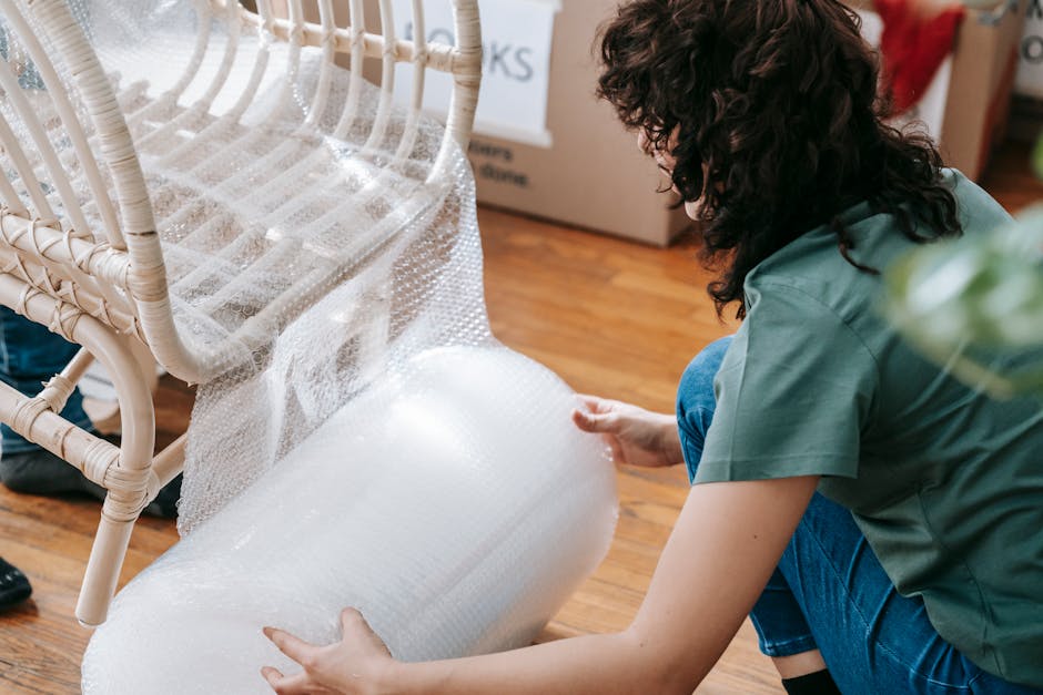 A woman and a man are engaged in surface cleaning and protective measures during a furniture move in a bright, well-lit living room. The woman is holding a roll of bubble wrap, preparing to protect a wooden chair with a fabric backrest, while the man is demonstrating careful handling of the chair’s seat. The room features a polished wooden floor, clean and shiny with no visible dust or debris. In the background, there are large windows with sheer white curtains, allowing natural light to fill the space, and a tall metal shelving unit with potted plants and decorative items. Cardboard boxes and a cardboard sleeve are also visible, indicating ongoing moving or rearrangement activity. This scene exemplifies proper techniques for protecting floor finishes and surfaces during furniture relocation, reflecting the importance of careful preparation in domestic cleaning and maintenance, as recommended by iCarpet Cleaners' guide to protecting floor finishes during furniture moves.